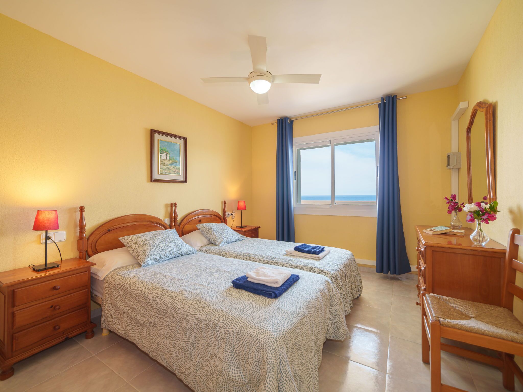 A hotel room with two beds, a ceiling fan, a window with curtains, a desk with a mirror, and a dresser.
