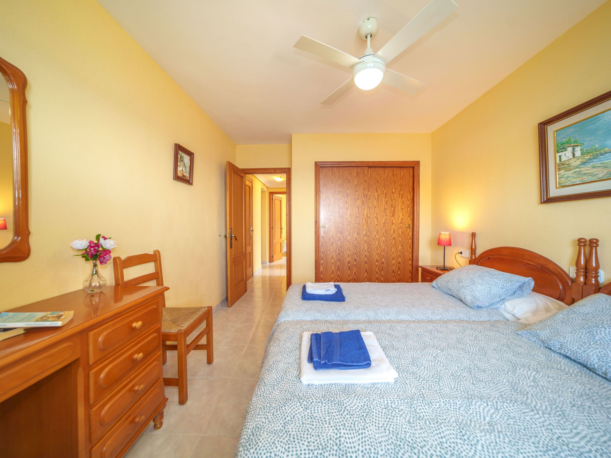 Habitación de hotel con dos camas, un escritorio, una silla, un aparador, ventilador de techo y un cuadro en la pared.