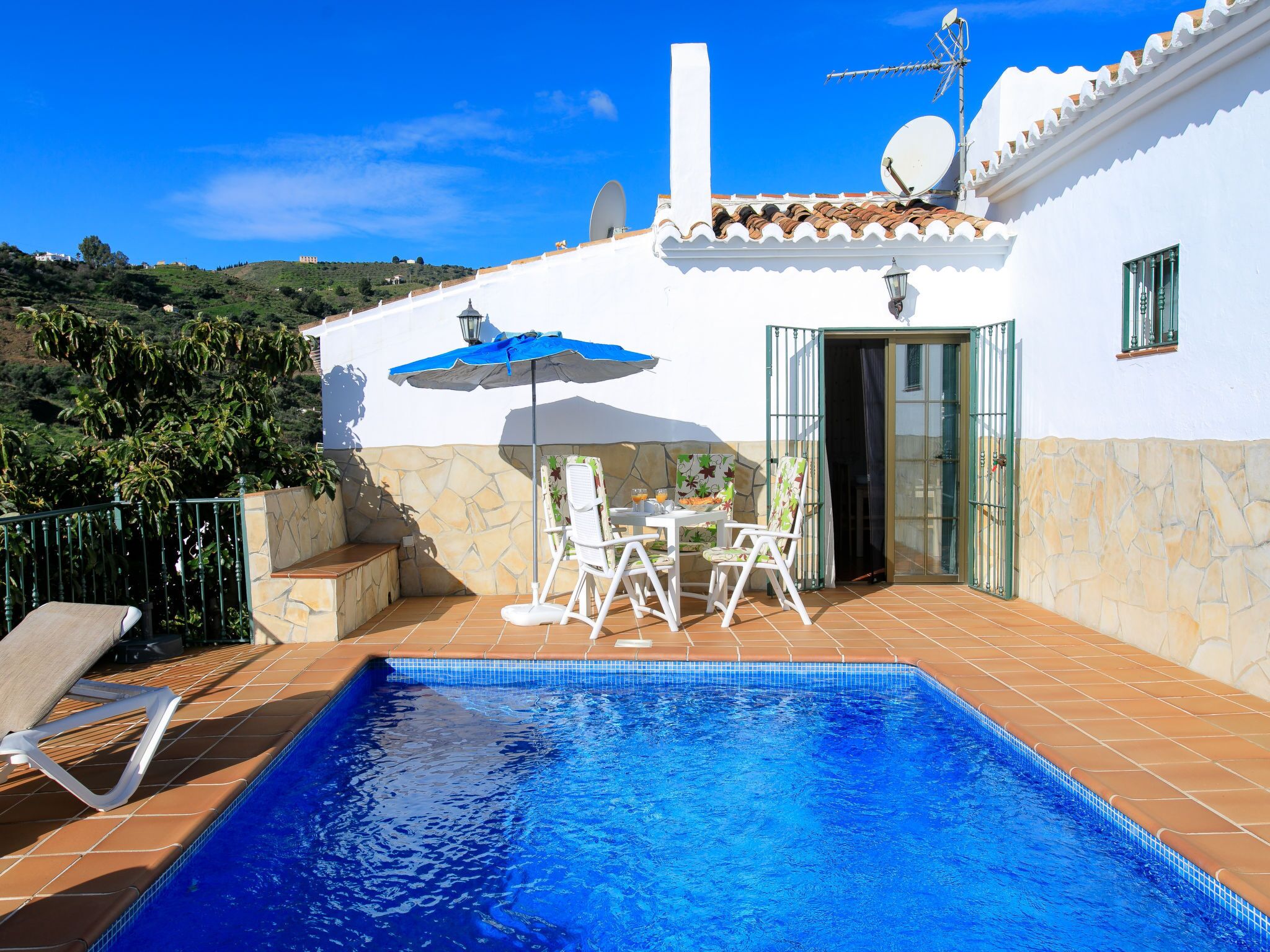A swimming pool with a tiled deck, a table with chairs, and a blue umbrella.