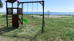 Un conjunto de columpios de madera con un pequeño refugio, un columpio verde y uno rojo, y una zona cubierta de césped con una playa al fondo.
