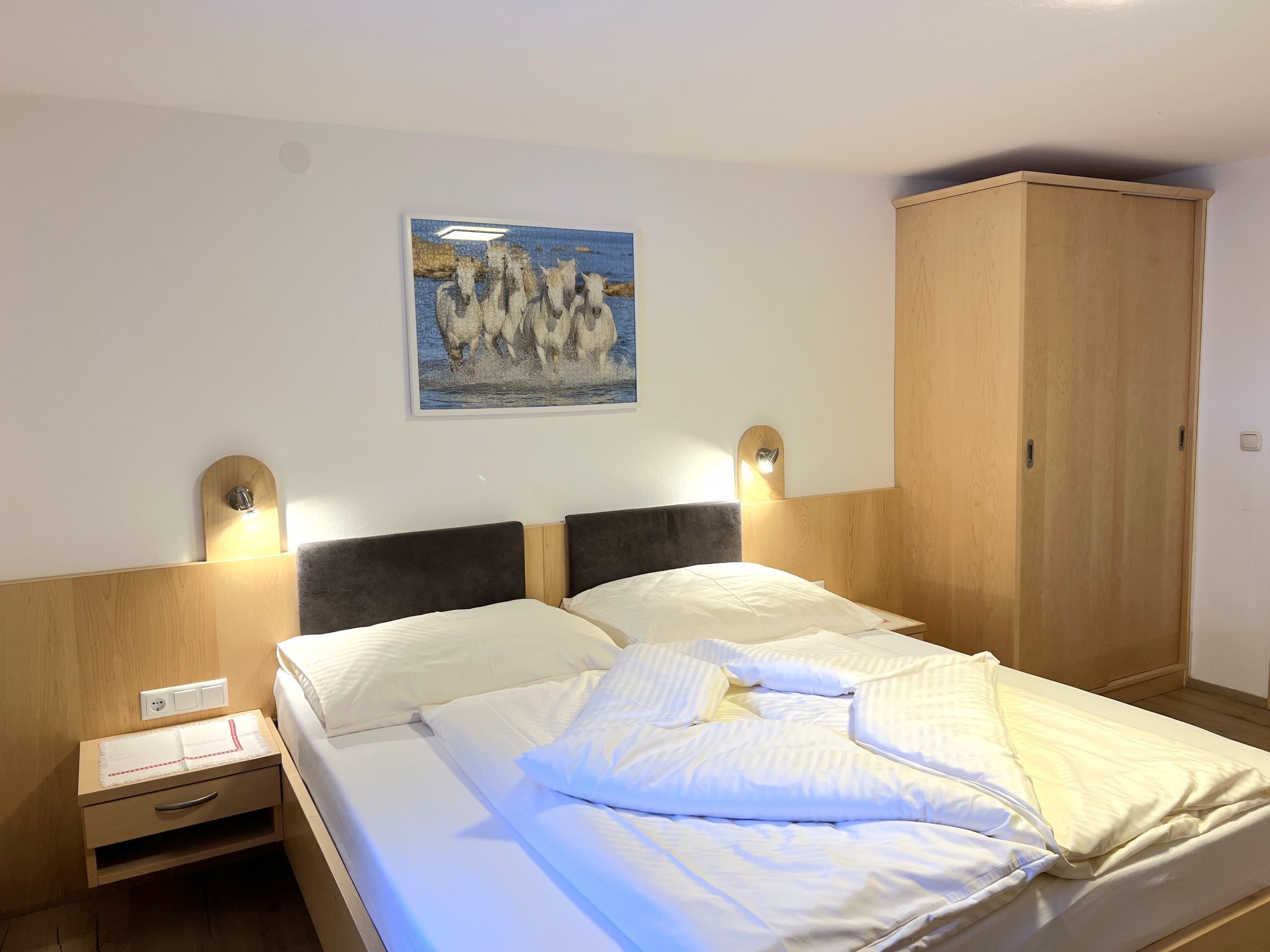 A neatly made bed with white linens, a wooden headboard, and a wall-mounted painting of horses.