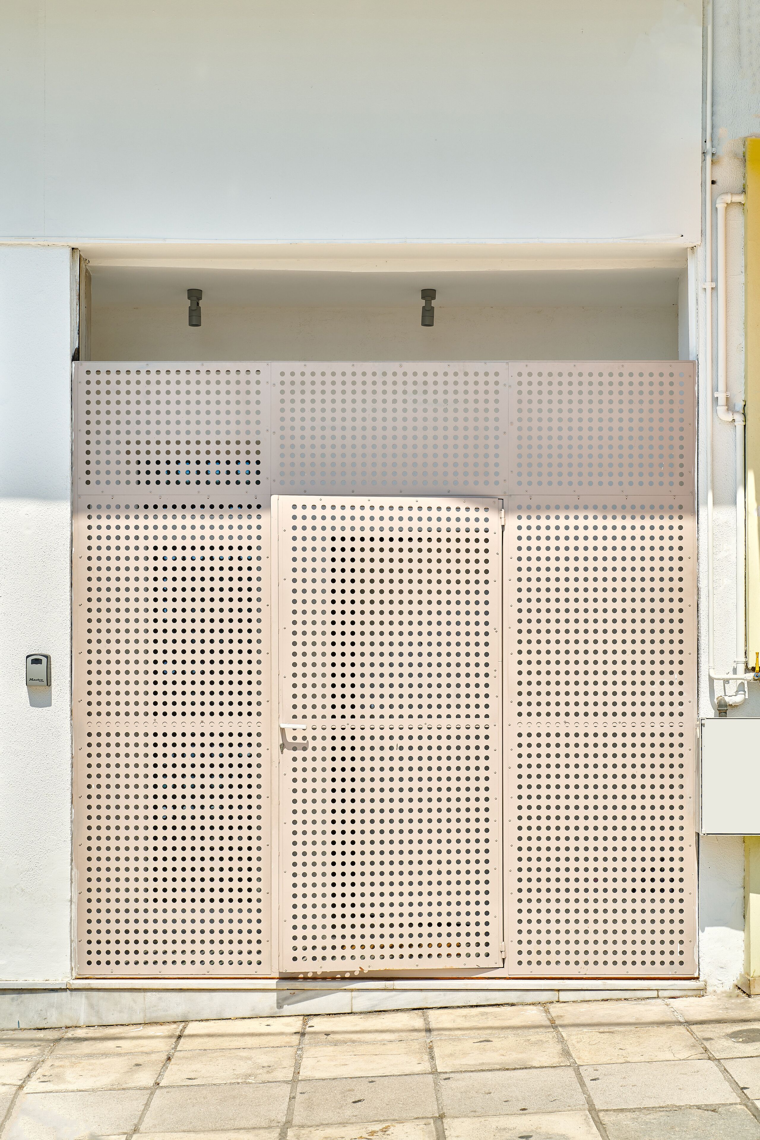 Une porte en métal, ornée d’un motif ajouré, est fixée à un bâtiment dont le mur est blanc et le sol recouvert de carreaux.