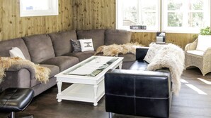 Un salon chaleureux avec un canapé d’angle gris, une table basse blanche et un pouf en cuir noir. On y trouve également un fauteuil en osier et une petite table d’appoint avec une lampe.