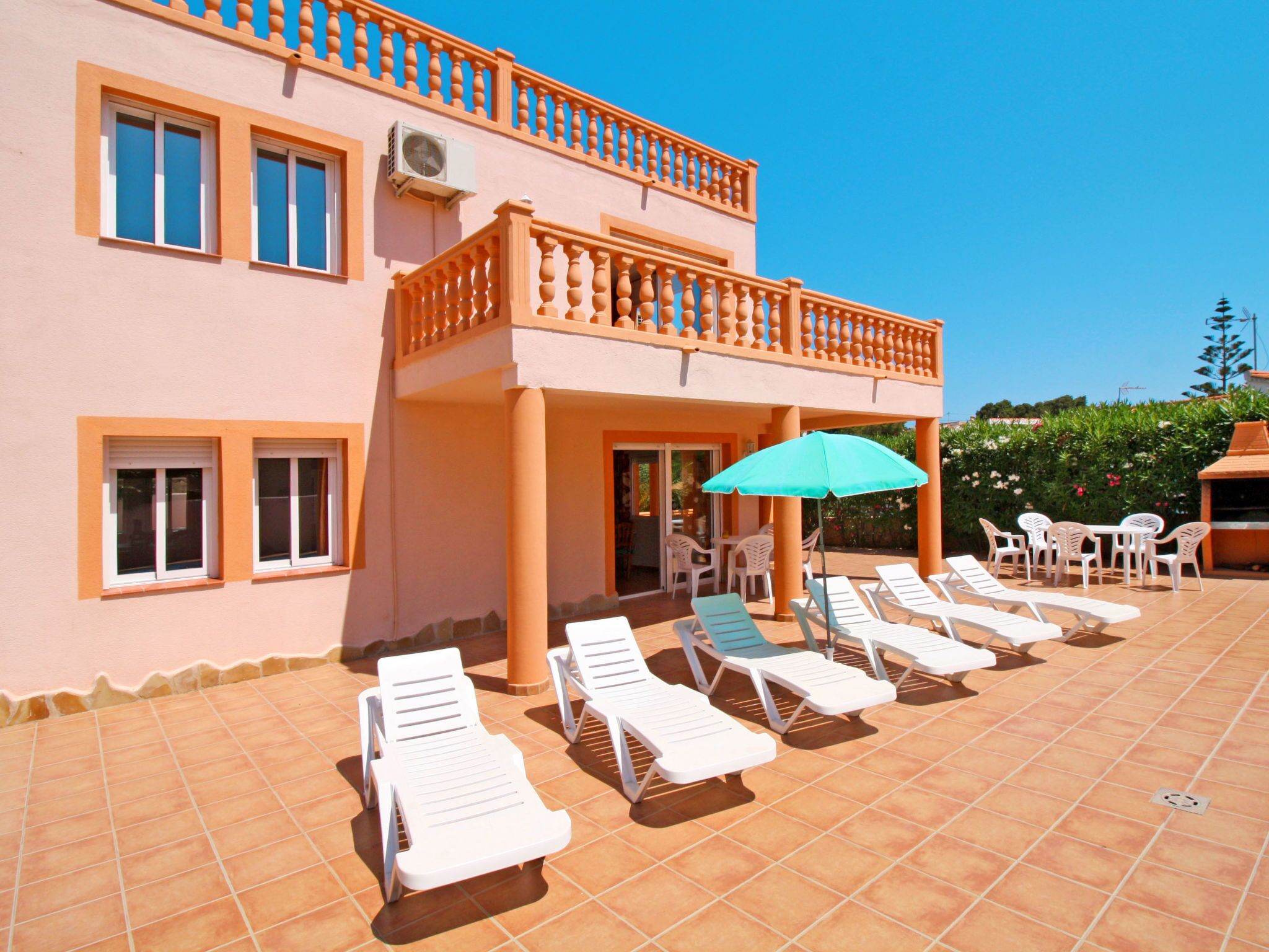 Ein zweistöckiges Gebäude mit Balkon, Außenbereich mit weißen Lounge-Stühlen und einem Tisch sowie einem grünen Sonnenschirm.