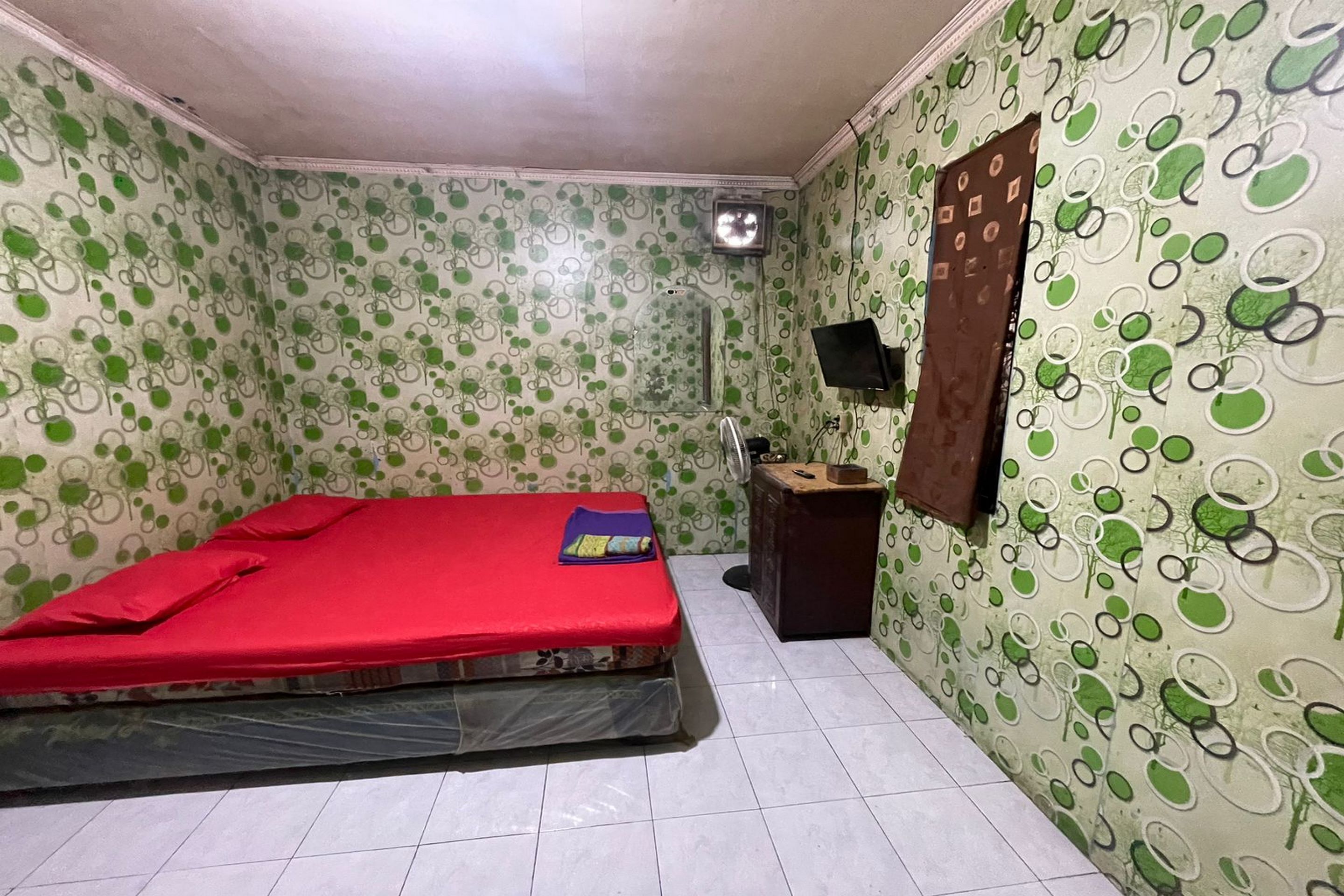 A small, single-bed room with a red mattress, a small bedside table, a wall-mounted TV, and patterned wallpaper.