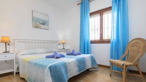 Ein Schlafzimmer mit einem Bett, zwei Nachttischlampen, einem Korbstuhl, einem Fenster mit blauen Vorhängen und einem Bild an der Wand.