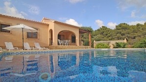 Ein Schwimmbecken mit gefliestem Rand, eine Lounge mit weißen Stühlen und einem Tisch sowie ein Gebäude mit Bögen und einem Balkon.