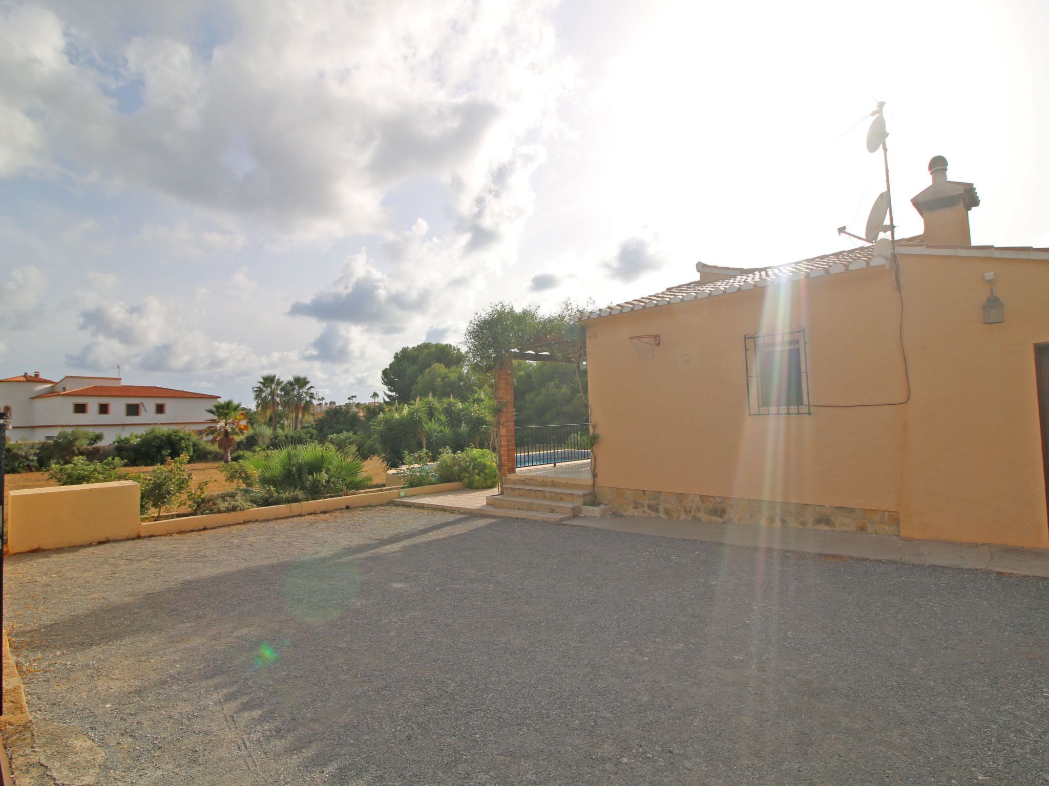 A residential area with a paved driveway, a small building with a satellite dish, and a white house in the background.