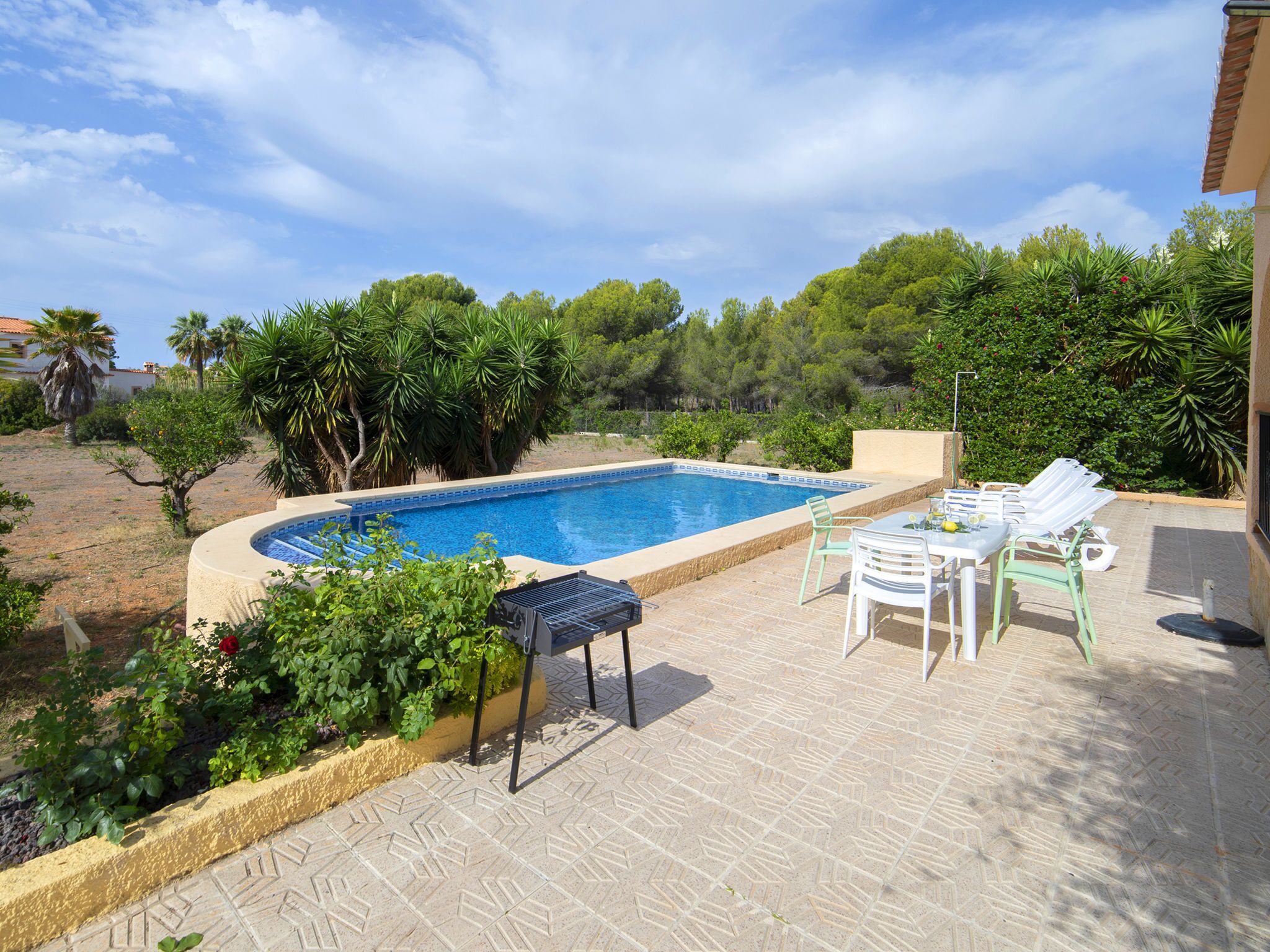 A swimming pool with a tiled deck, surrounded by greenery and a few chairs.