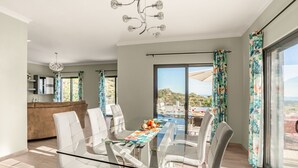 Un comedor moderno con una mesa de vidrio y sillas blancas, una cocina al fondo y un amplio ventanal con vistas al exterior.