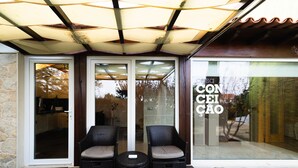Un espace extérieur abrité avec deux fauteuils en osier et une petite table ronde, un mur en pierre et une porte vitrée donnant sur un patio.