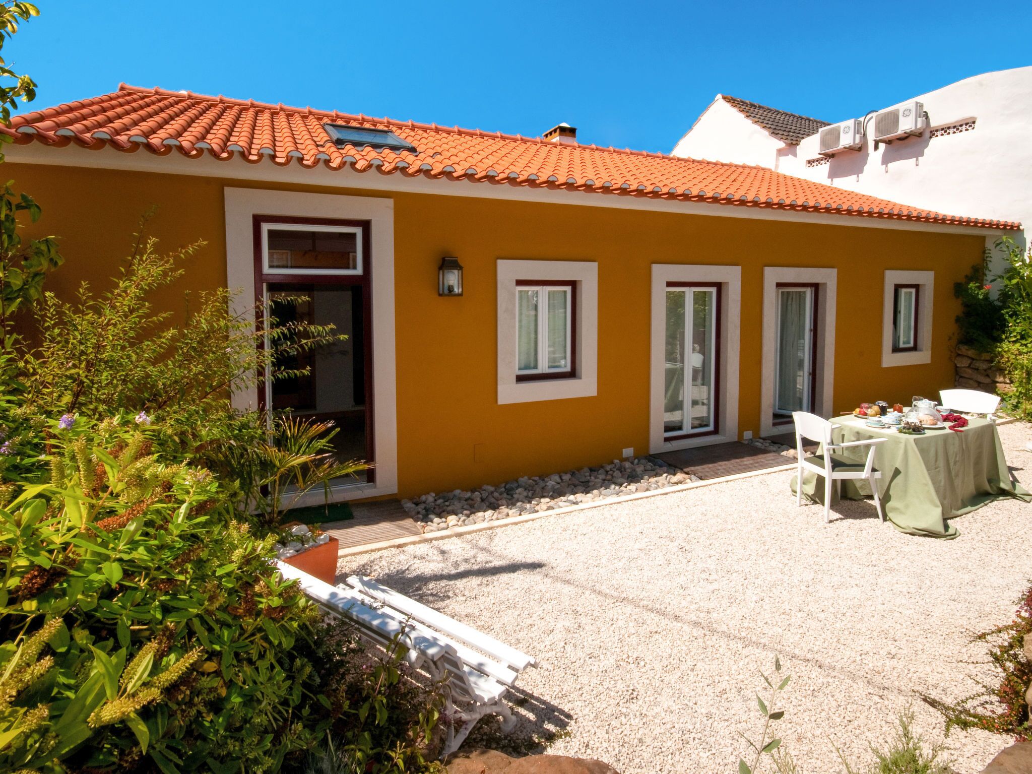 Un bâtiment jaune avec un toit recouvert de tuiles rouges, une terrasse avec une table et des chaises, et un sol en gravier.