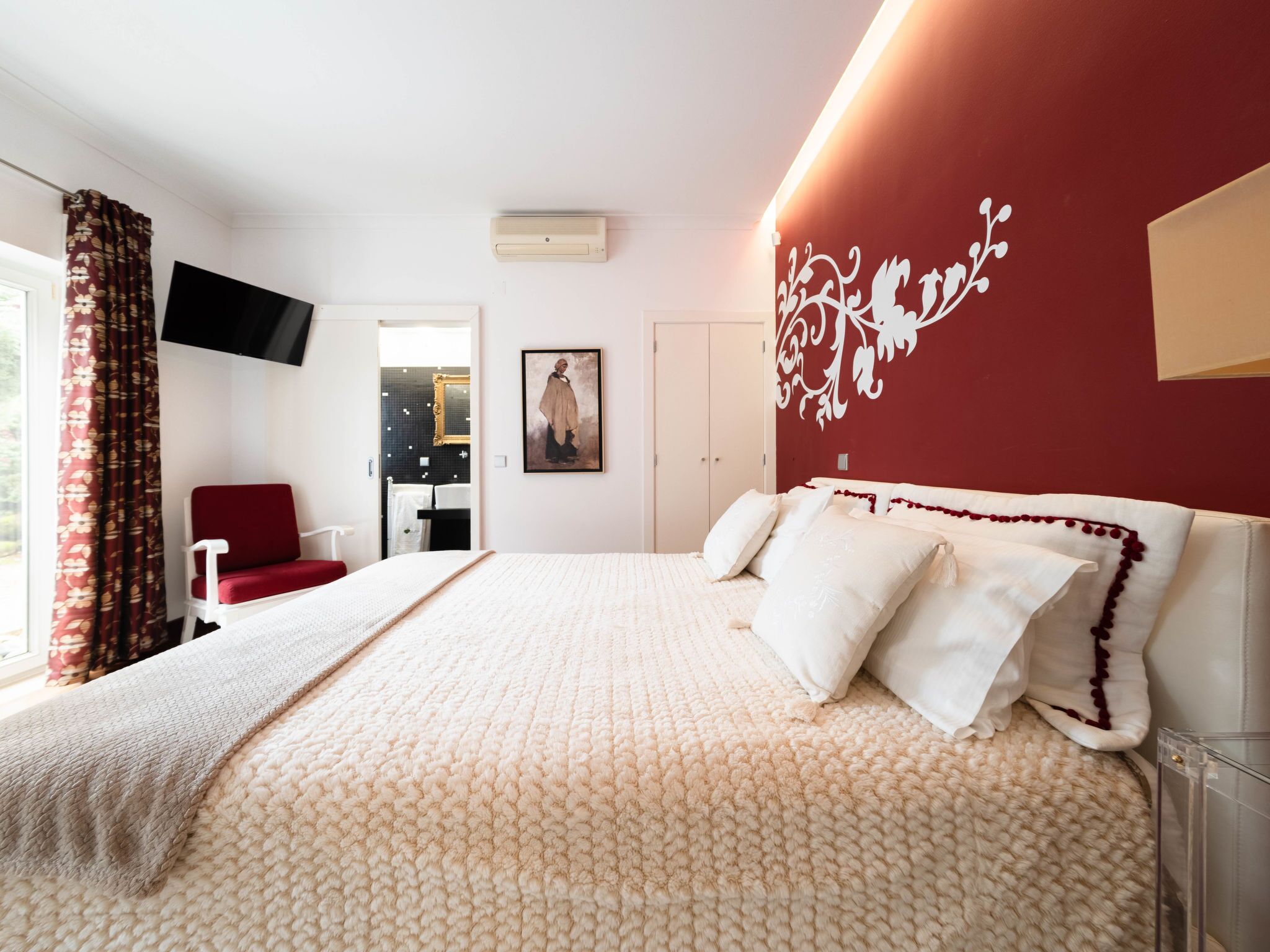 A bedroom with a large bed, a red chair, a TV mounted on the wall, and a bathroom visible through an open door.