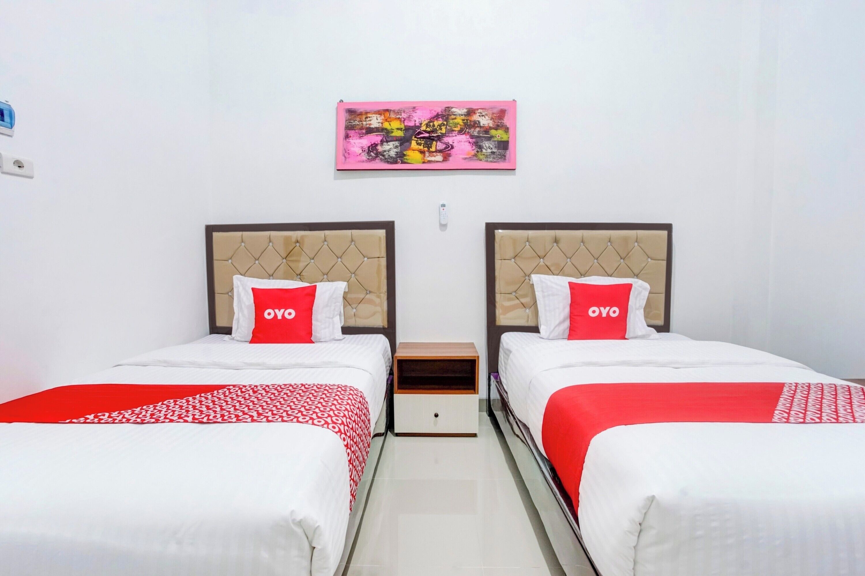 Habitación de hotel con dos camas, cada una con cubrecama rojo y blanco y almohadas a juego. Hay una mesita de noche de madera entre las camas, y un cuadro abstracto rosa cuelga en la pared de arriba.