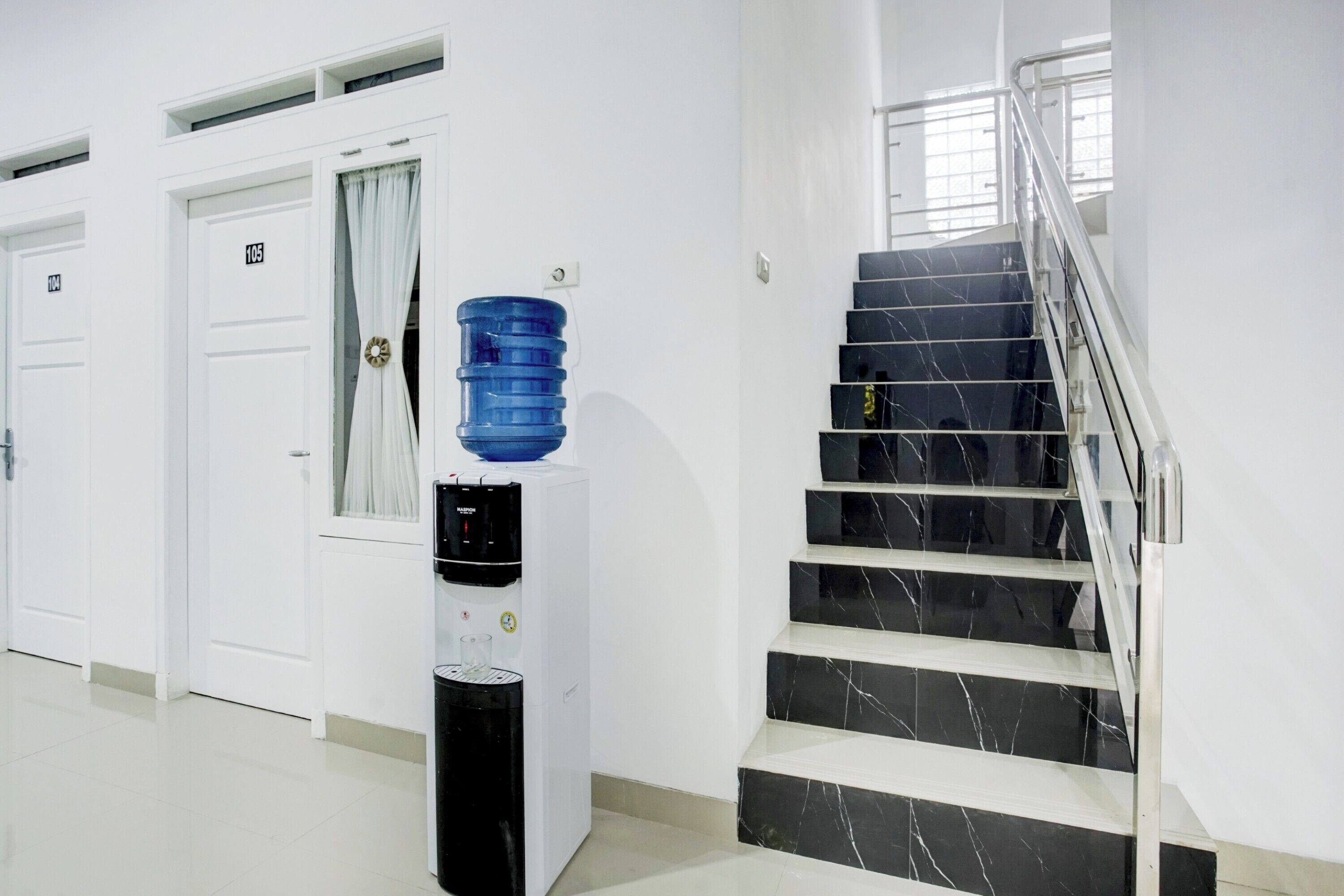 A hotel corridor with a water dispenser, a staircase with black marble steps, and white doors numbered 100 and 105.