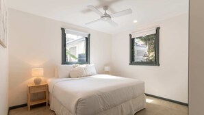 Une chambre à coucher comprenant un lit, une table de chevet, une lampe, un ventilateur de plafond et deux fenêtres.