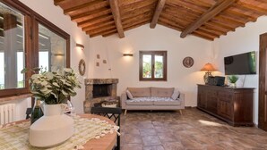 Una sala de estar acogedora con chimenea de piedra, vigas de madera en el techo, un sofá, un televisor y una mesa de comedor con un jarrón de flores.