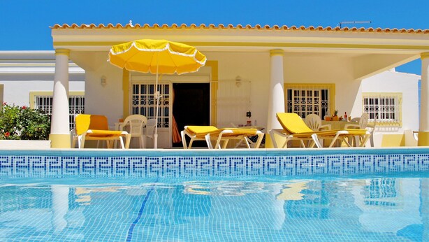 Un espace aménagé au bord de la piscine, avec des chaises jaunes et un parasol, une piscine carrelée et un bâtiment aux murs blancs et aux bordures jaunes.