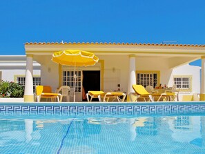 Un'area piscina con sedie gialle e un ombrellone, una piscina piastrellata e un edificio con pareti bianche e dettagli gialli.