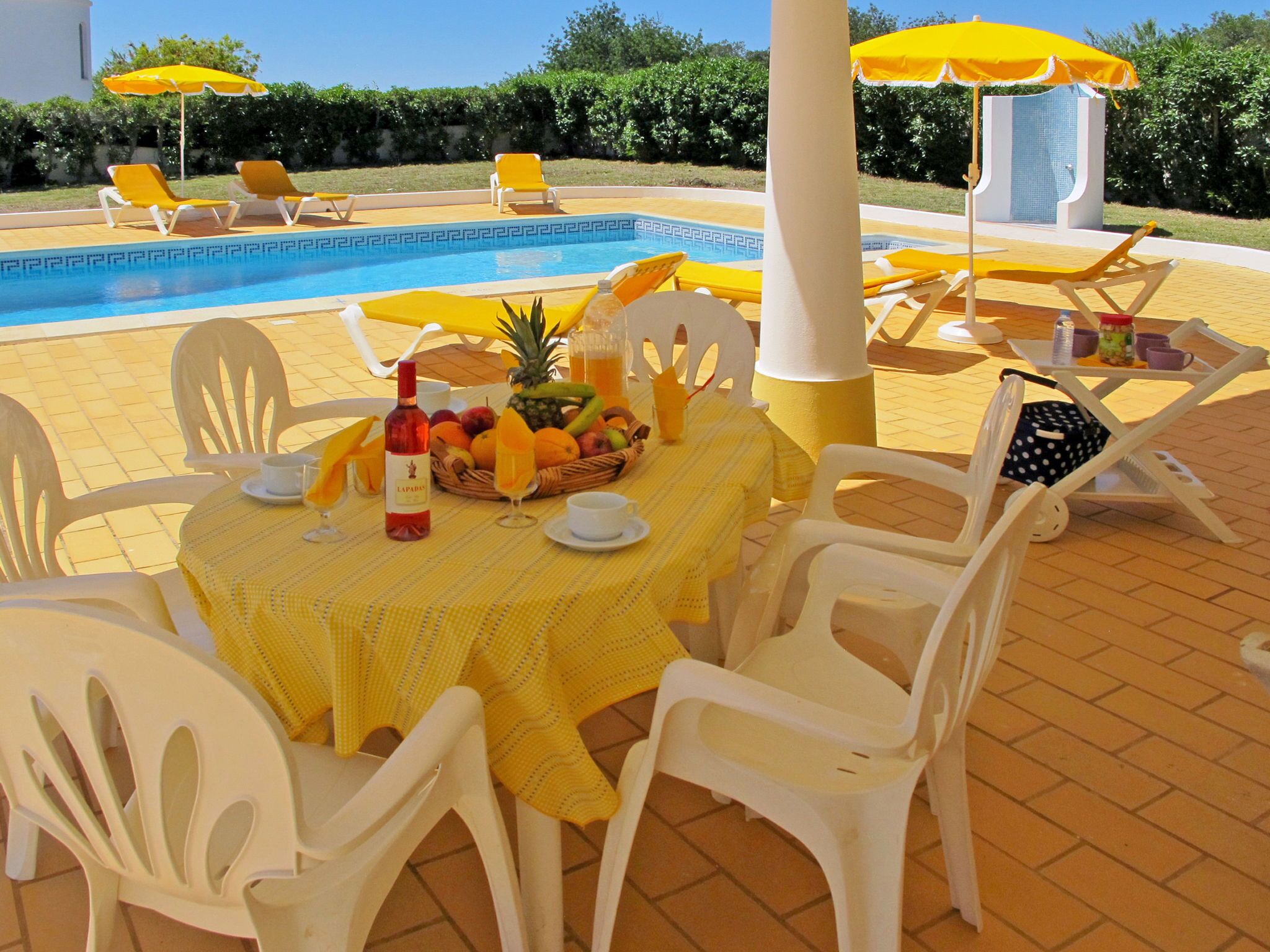 Un área de comedor junto a la piscina con sillas de plástico blanco y sombrillas amarillas, una mesa dispuesta con frutas, una botella y vasos.
