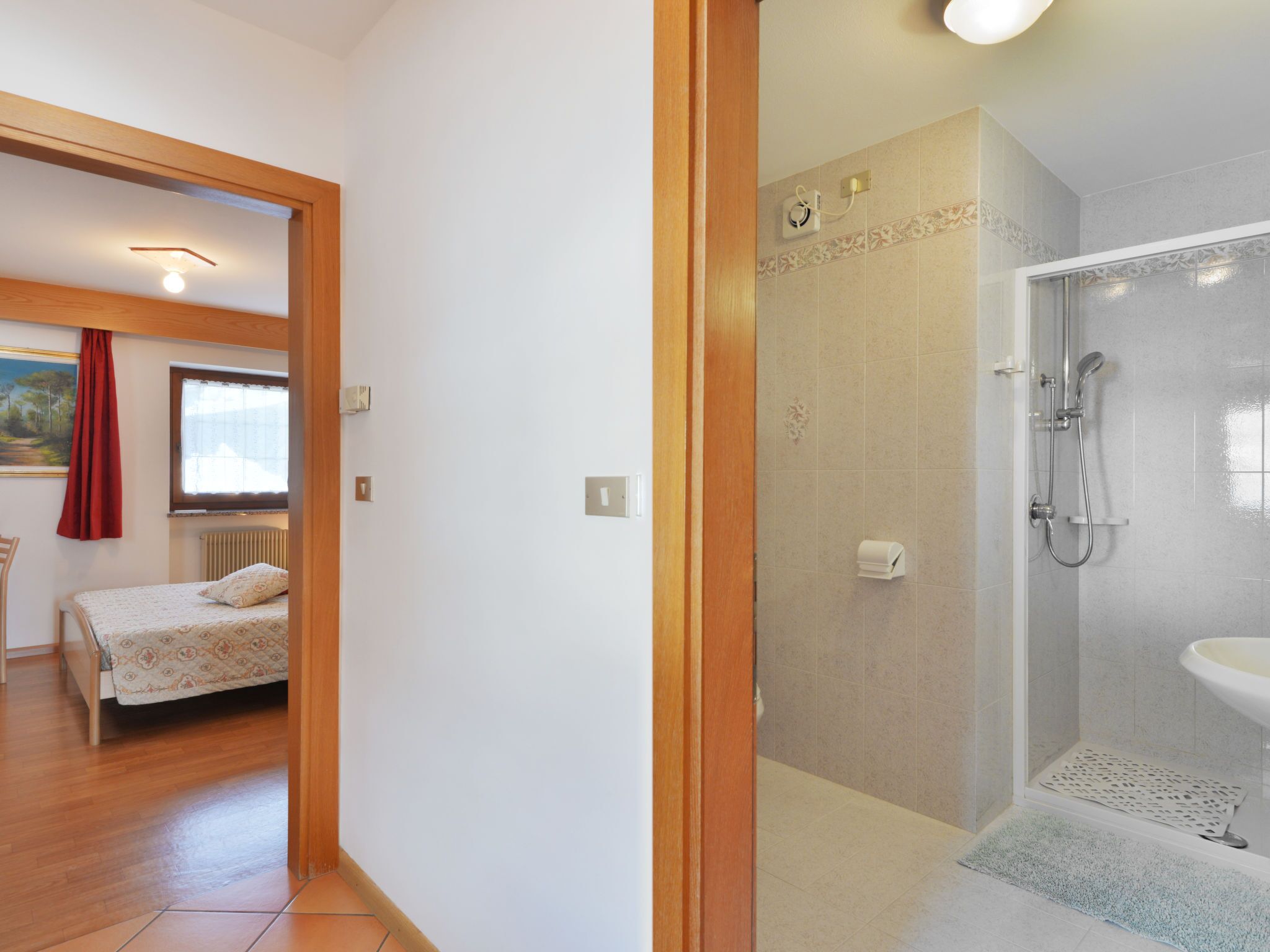 Quarto com cama, janela com cortinas, radiador e porta que dá para um banheiro com chuveiro e pia.