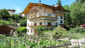 A multi-story building with balconies adorned with flowers, surrounded by lush greenery and a wooden fence. - Pancheri by Interhome (Valle di Non)