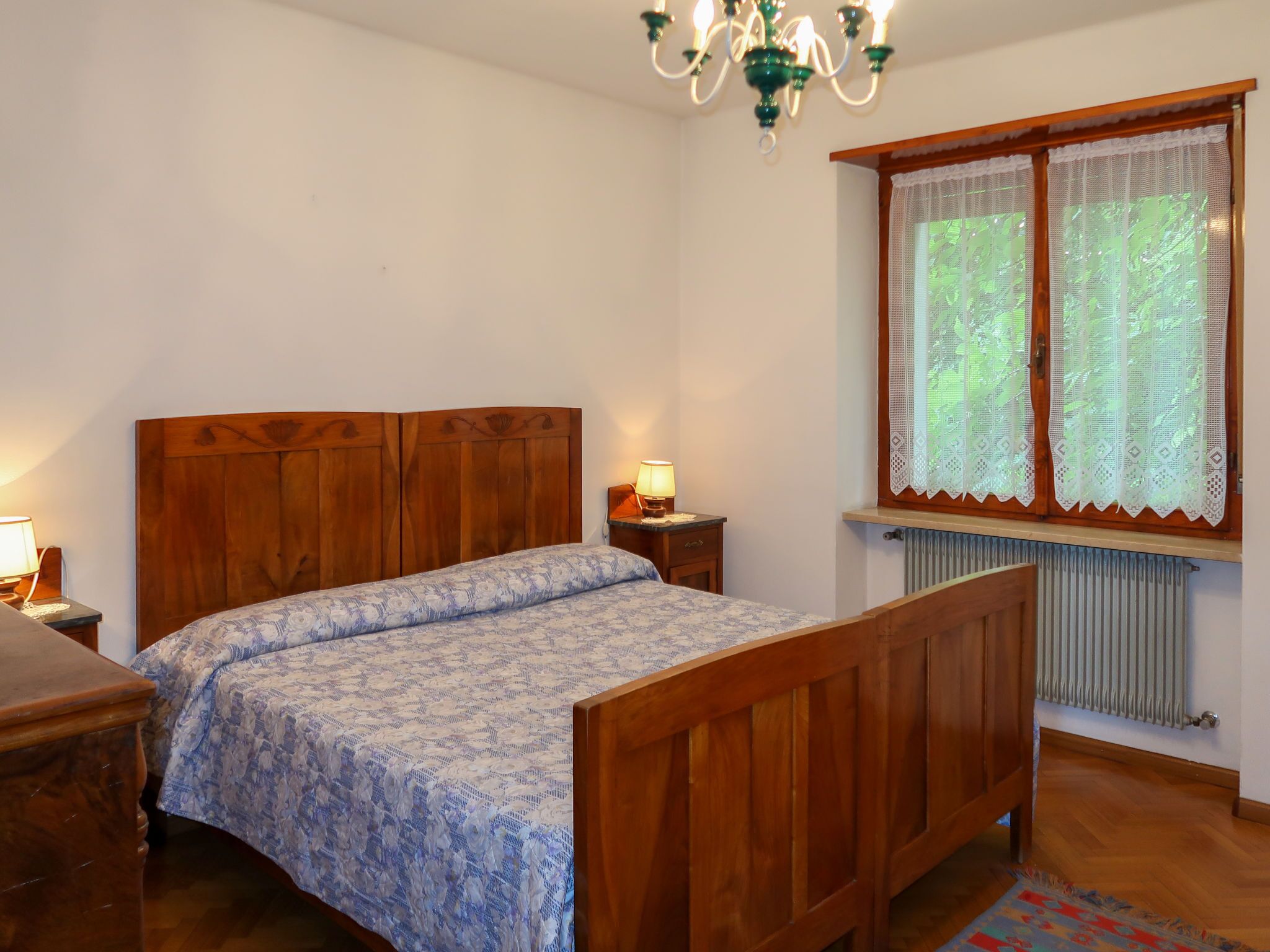 Et soveværelse med en træseng, et natbord med en lampe, et vindue med blondegardiner og en radiator.
