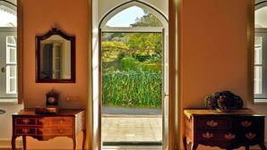 A room with a large arched doorway leading to a garden, two wooden side tables with drawers, a mirror on the wall, and a striped armchair.