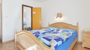 A bedroom with a wooden bed, a bedside table, a nightstand, a wall-mounted light fixture, and a door leading to another room.