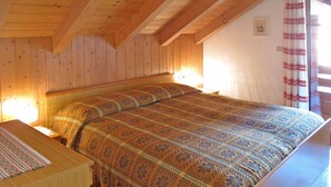 Een slaapkamer met een houten plafond, een bed met een beddengoed met dessin, een nachtkastje en een raam met gordijnen.