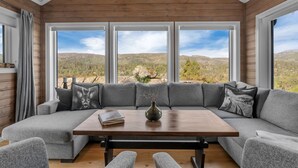 Een moderne woonkamer met een houten plafond, grote ramen, een hoekbank, een salontafel en uitzicht op een bergachtig landschap.