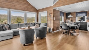 Een ruime woonkamer met een grote grijze bank, een eettafel en een keuken met houten kasten en uitzicht op de bergen.