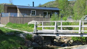 A wooden cabin with a metal roof, a bridge over a stream, and a forested hillside in the background.