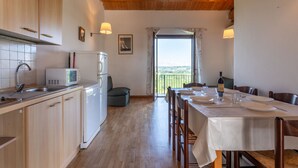 Een keuken met houten kasten, een magnetron en een koelkast. Er is een eettafel met stoelen en uitzicht op het platteland door een raam.