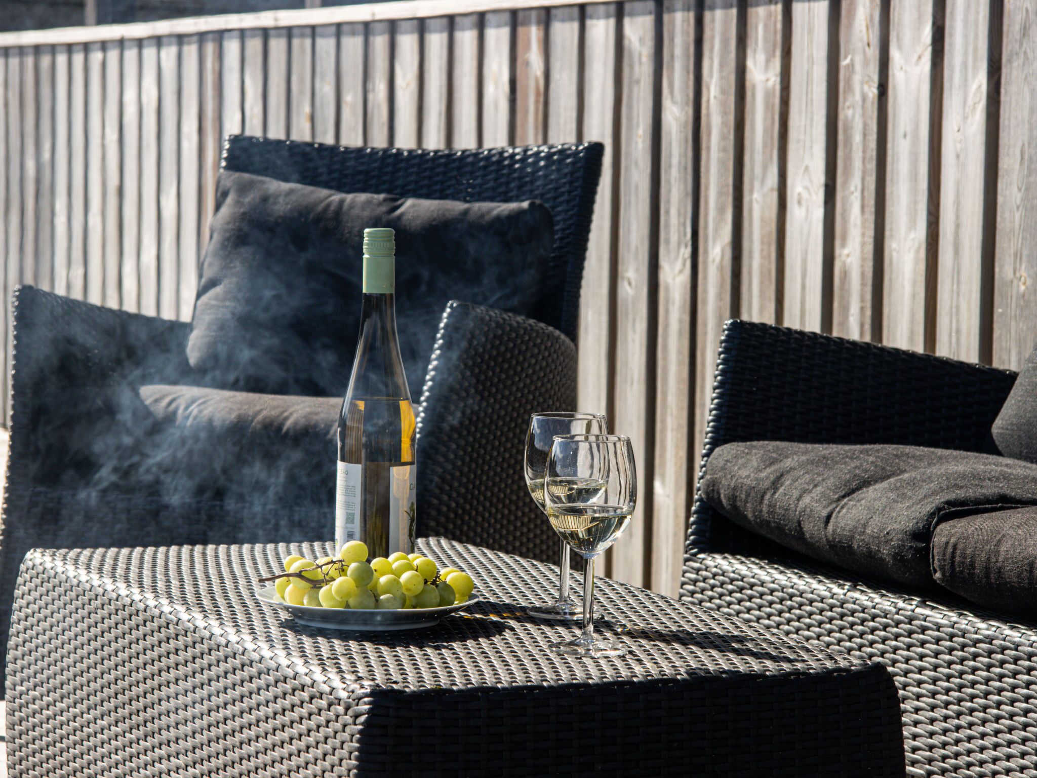 A wicker table with a bottle of wine, two glasses, and a bowl of grapes.