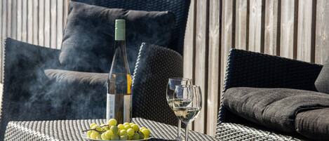 A wicker table with a bottle of wine, two glasses, and a bowl of grapes.