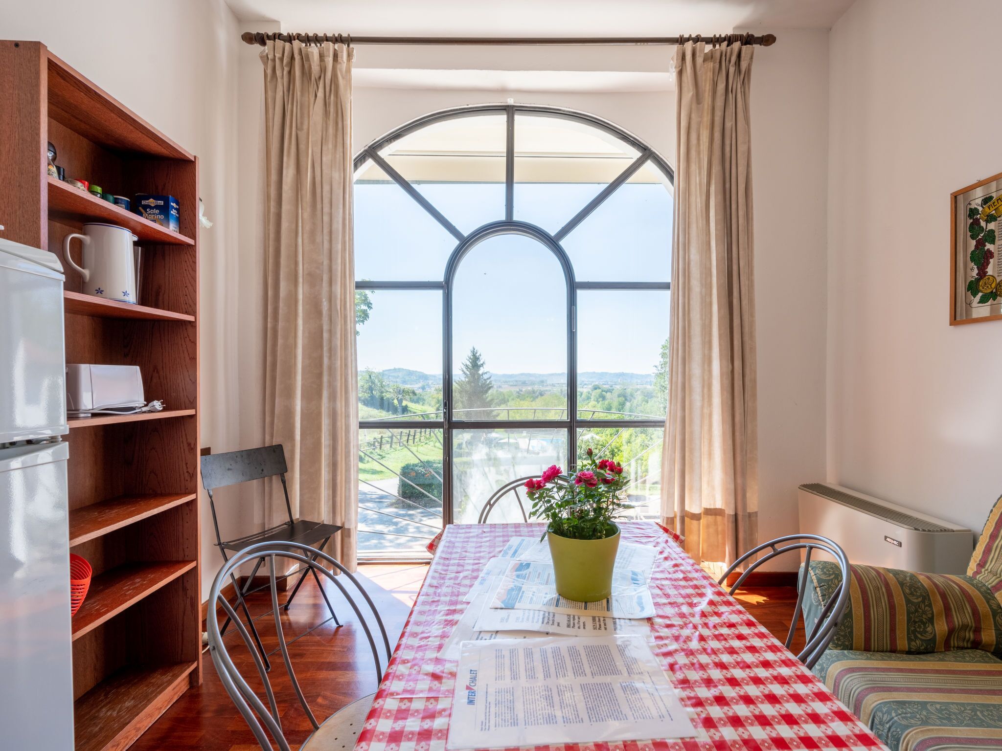 A dining area with a table set for two, a bookshelf, and a view through a large arched window.