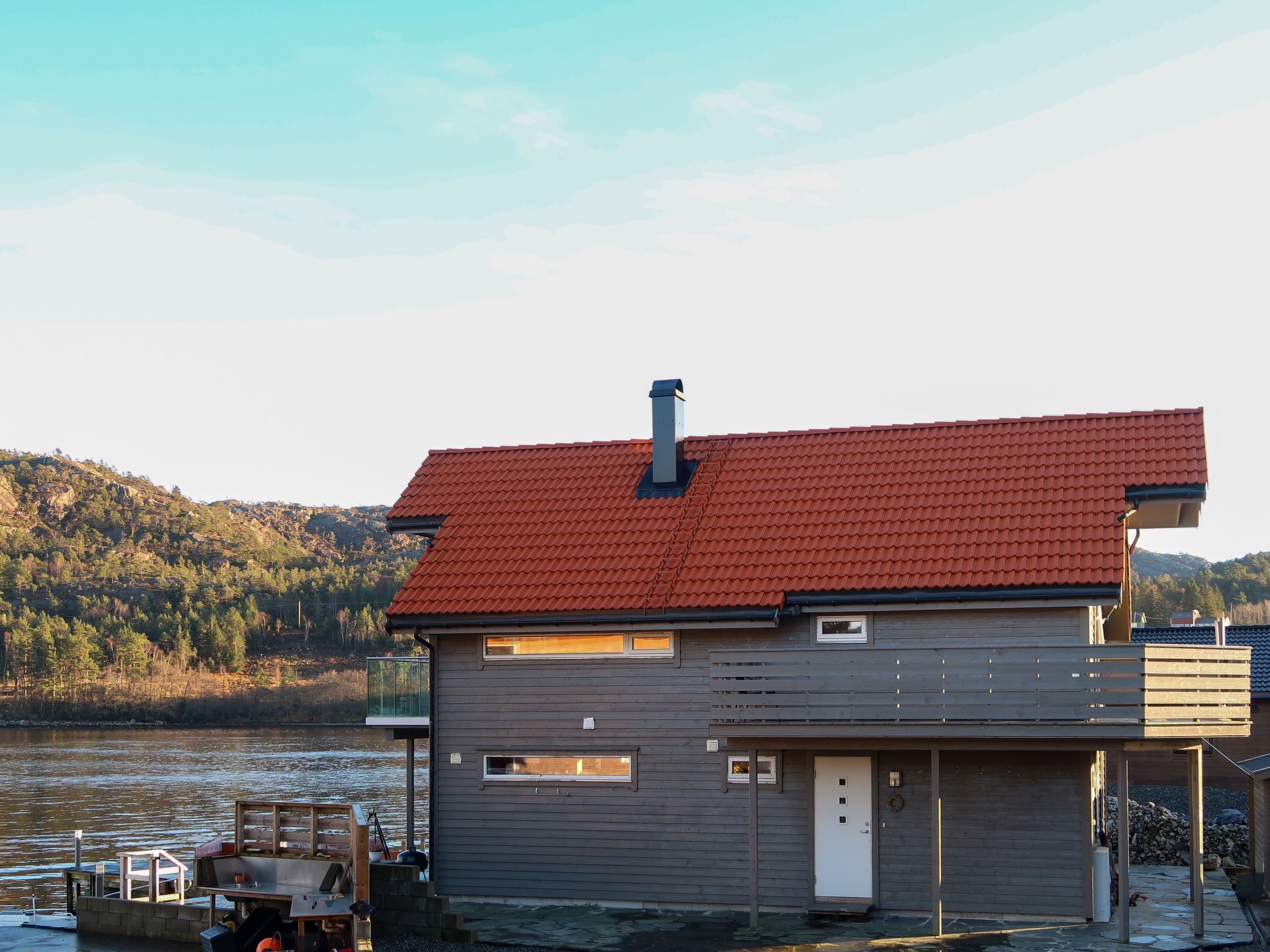Ein modernes Haus mit rotem Dach, einem Steg und Blick auf einen See sowie Hügel im Hintergrund.