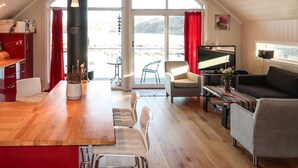 A spacious living area with a wooden dining table, a sofa, and a TV. There is a sliding glass door leading to a balcony with a view of the outdoors. - Jektastøo by Interhome (Huglo)