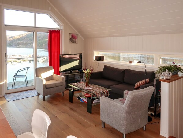 Una sala de estar con sofá, sillones, una mesa de centro, televisor y vista a una masa de agua a través de amplias ventanas.