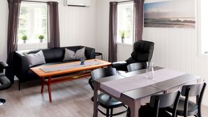 A modern room with a black leather sofa, a wooden dining table, and a black leather chair. There is a window with curtains, a wall-mounted air conditioner, and a painting of a landscape. - Nesholmen by Interhome (Leirvik)