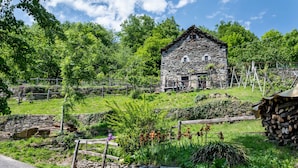 A stone building with a thatched roof, surrounded by lush greenery and a stone wall. - Baita Degli Orsi by Interhome (Domodossola)