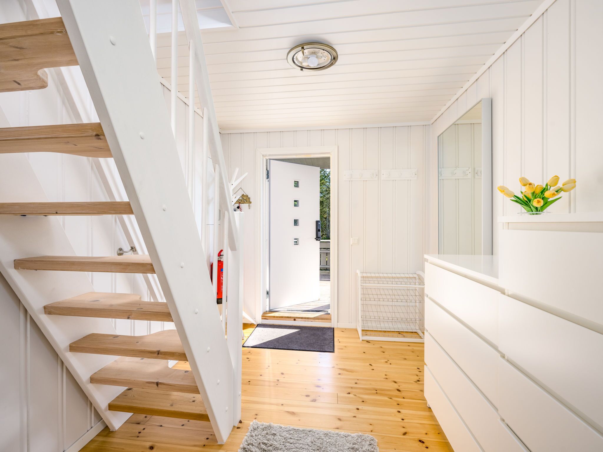 Un intérieur moderne avec un escalier en bois, des murs blancs et une vue sur une pièce où se trouve un paillasson noir à l’entrée.
