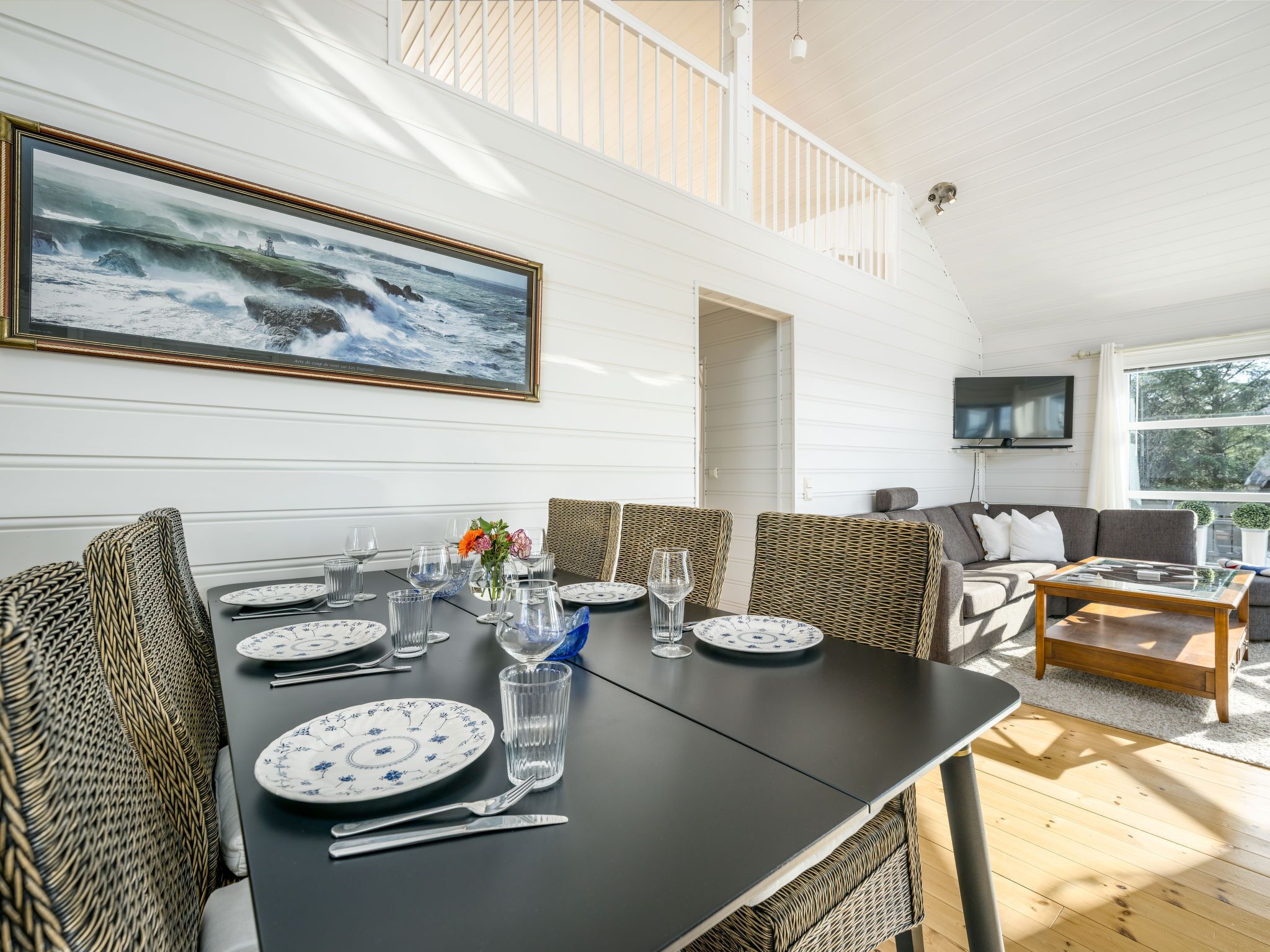 A dining area with a black table set for six, surrounded by wicker chairs, a wicker sofa, and a wooden coffee table. A large framed picture of a coastal scene hangs above the table.