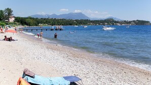 Una playa de guijarros con gente tomando el sol, un muelle que se adentra en el agua y barcos al fondo.