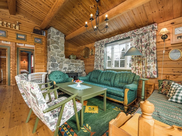 A cozy cabin interior with a stone fireplace, wooden walls, a green sofa, floral-patterned chairs, and a wooden dining table. - Fjellro by Interhome (Haukedalsvatn)