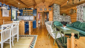 Een gezellig houten interieur met een houten plafond, een stenen open haard, een eethoek met witte stoelen, een groene bank en een keuken met blauwe en witte tegels.