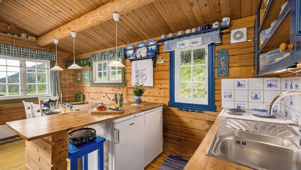 Een houten blokhut van binnen met een keuken, eethoek en een raam met uitzicht op groen.