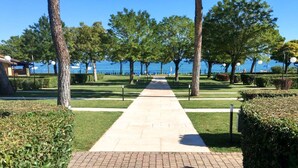 A paved pathway leading to a body of water, flanked by trees and well-maintained hedges.