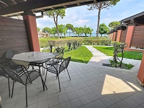 Un patio al aire libre con una mesa redonda y sillas, piso de baldosas y vistas a un jardín ajardinado con un camino y árboles.